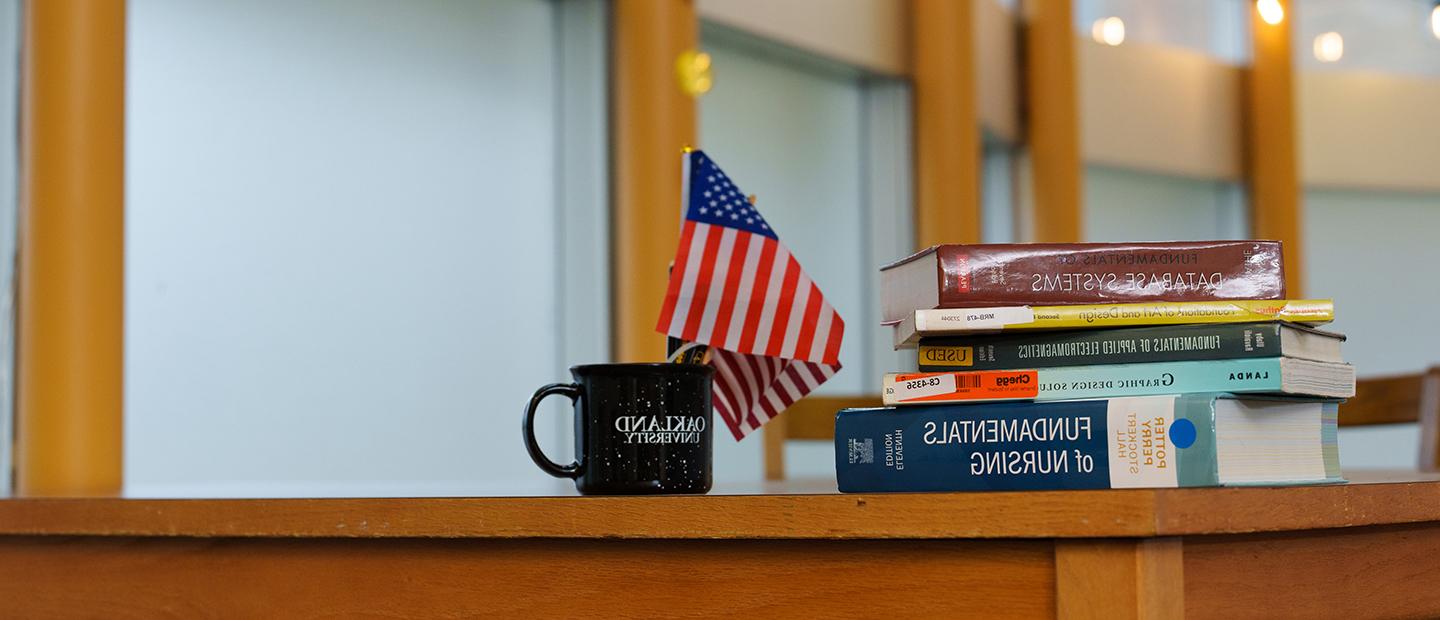 A pile of text books sits on a desk along with an Oakland University coffee mug that contains small American flags.